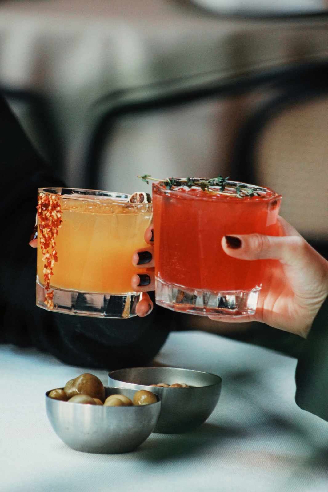 Two cocktails raised in a toast, amber and red, olives on the table, warm light.
